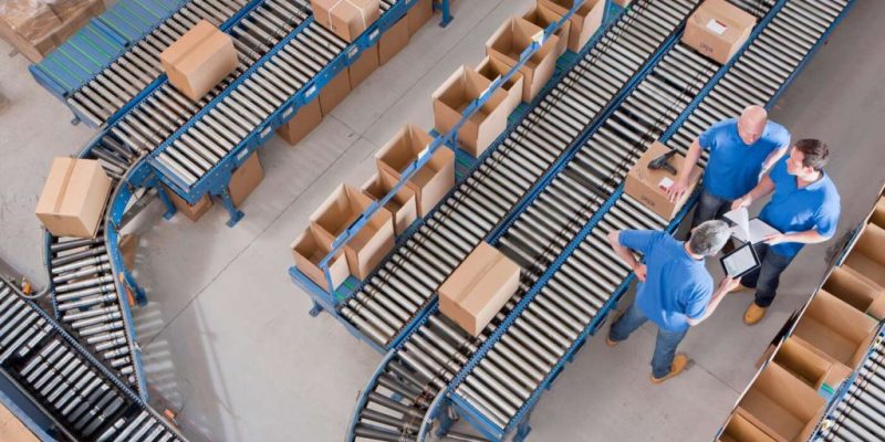Top view of workers with papers and a digital tablet having a discussion among boxes laid on conveyor belts at a distribution warehouse.
